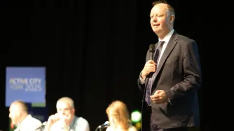 A man in a dark suit wearing a tie and holding a microphone stood on a stage in front of a table of delegates.