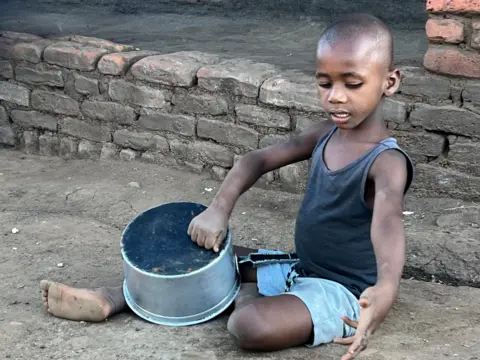 A young boy sits on the ground in shorts and a vest playing with a cooking pot.