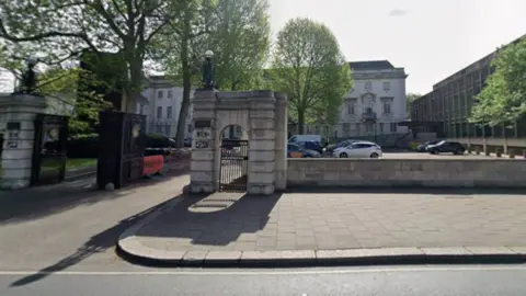 Google A court building with a car park in front of it. There is an arched pedestrian entrance and a gated vehicle entrance. There are a number of vehicles parked in front of the building. There is a pavement and a road running alongside a a wall which bounds the car park.