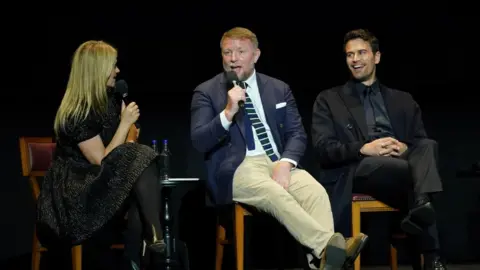 PA Media Guy Ritchie (centre) and Theo James (right), speaking to Edith Bowman. They are sat on chairs and Guy Ritchie is talking into a microphone. There is a black background behind them.