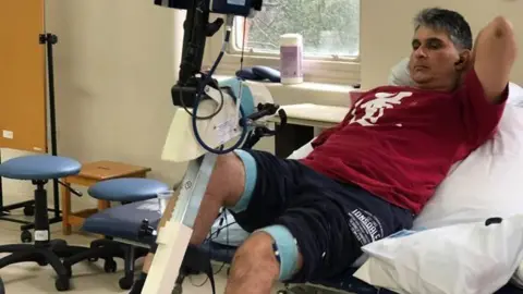 Niraj Haria in a red t-shirt on a hospital bed with his leg attached to a metal rod with wires and monitoring equipment