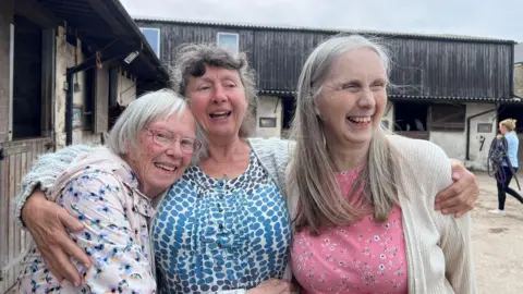 The Rainbow Care Group Three women with silver hair smile at the camera and hug each other. They stand in a yard near some stables.