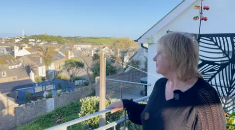 A woman with a black long sleeve top and a short blond bob looks out over railings to a view of housing and green fields in St Mary's on a sunny day with clear blue skies.