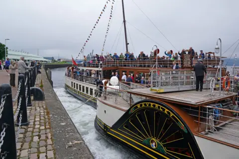 PA Media A paddle steamer filled with people, with some waving at the those on the dock.