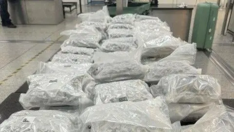 NCA Lots of vacuum sealed white plastic bags laid out on a table. Two green suitcases are in the background in an airport hall.
