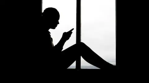 A high-contrast silhouette of a person sitting indoors in front of a large window. The person is positioned in profile view, facing toward the left side of the image. The outline of the figure is entirely dark, with no facial details visible due to the strong backlighting from the window behind.