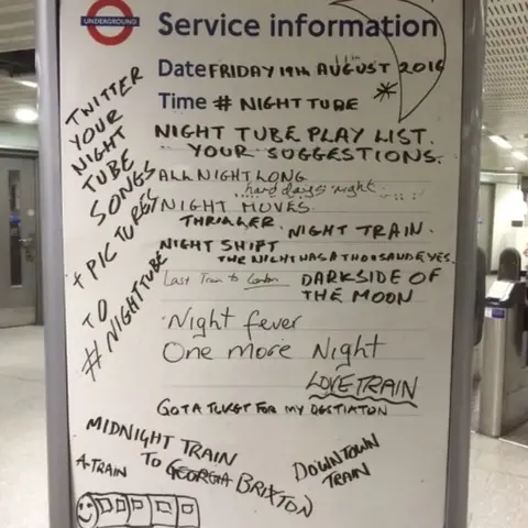 A London Underground service information board covered with handwritten song suggestions for a “Night Tube Playlist.” The board includes titles such as “Night Fever,” “One More Night,” “Love Train,” and “Dark Side of the Moon,” along with doodles and notes from passengers.