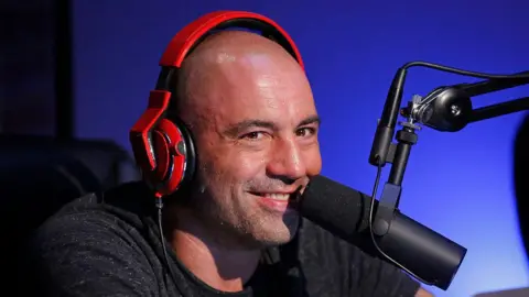 Getty Images Joe Rogan, wearing orange headphones, smiles into a microphone during an episode of Joe Rogan Questions Everything