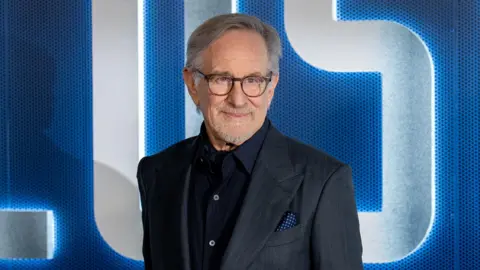 Reuters Steven Spielberg looks off camera. He is smiling and wearing a dark grey suit with dark blue open-necked shirt and a spotted blue handkerchief in his breast pocket.