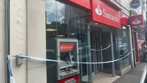 The front of a bank branch with red and white signage including the Santander logo and name. The whole frontage is festooned with blue and white police tape. A cash machine is visible but not obviously damaged from the front.