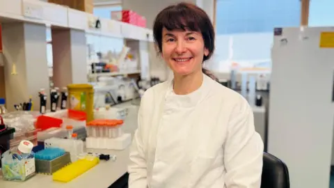 Professor Dana Dawson smiling at the camera, she is in a laboratory setting wearing a white clinical jacket.