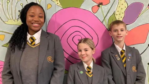 Three children in grey school uniforms with yellow and black ties pose, smiling, shoulder to shoulder in front of a large mural inside Aureus School in Didcot