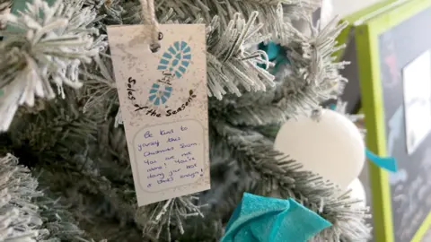 A message is pinned to an artificial, Christmas tree, alongside the more traditional baubles. It tells people "Be kind to yourself - you are not alone."