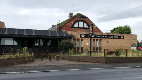 Terry Gormley Maltby Miners Welfare Institute was a brown and red-brick building with a large black and white sign across the entrance. A patch of long grass and wooden picnic benches can be see outside.