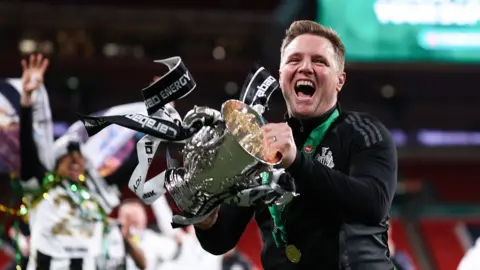 Jacques Feeney/Offside/Offside melalui Getty Images Eddie Howe di lapangan di Wembley, tersenyum memegang trofi Piala Carabao yang memiliki pita hitam dan putih menggantungnya.