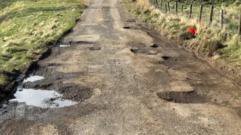 Harry Byatt A stretch of unclassified road with potholes in it and a fence to one side 