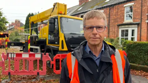 Andrew Wilson has short, light-brown hair and wears blue-rimmed glasses, a black jacket, a blue shirt and an orange and silver hi-vis vest. He stands in front of a yellow lorry, which is carrying a crane, and red-brick period homes in Westbourne Avenue, Hull. Red barriers block the pavement, and orange and brown leaves cover the ground.