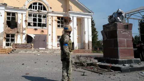 Shutterstock A Ukrainian soldier in military uniform with a blue armband stands in front of a heavily damaged building, its windows shattered and walls riddled with bullet holes, alongside the remnants of a destroyed Lenin statue in a war-torn urban area.