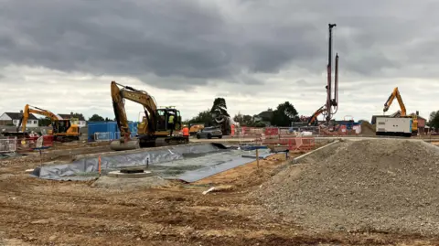 Owen Sennitt/BBC A number of diggers on a building site with upturned ground to prepare for the new building. It is a grey day. 