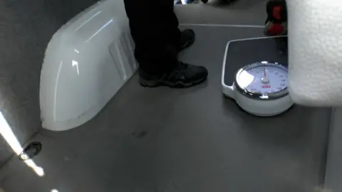 Swansea Trading Standards A set of black and white weighing scales sit on the dark grey floor at the back of a van. A white internal wheel arch is visible to the left of the frame. 