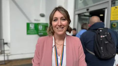 A women with short dark blonde hair smiling at the camera. She is wearing a pink blazer and a white blouse. She has a rainbow lanyard on.
