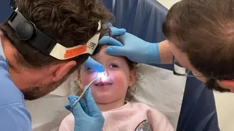 Peyton Handley - smiling and lying on a hospital bed as two doctors examine her nose, shining a torch and inserting tweezers in her nostril