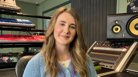 Vikki Irwin/BBC Megan Pilcher is sitting in the Level Two Charity recording studio. In the background there is a recording desk with multiple switches and buttons, a large speaker and keyboards. She is wearing a light blue cardigan with a light yellow top underneath.
