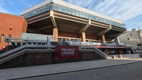 LDRS Image shows the late modernist-style Preston Guild Hall, built in 1973. It is a large concrete structure which expands outwards towards the top, reflecting the shape of the upper seating areas. 