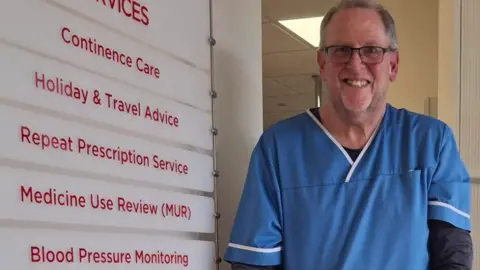 Mr Dugmore is wearing blue scrubs stood next a sign in the pharmacy.