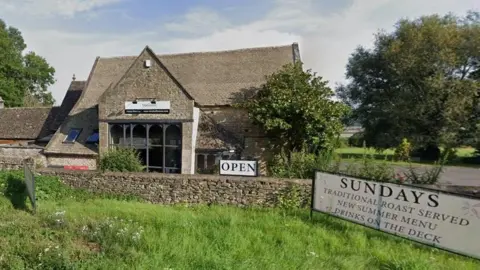 Google View of the pub from the road. It is a stone building with a sloping roof and a large floor-to-ceiling window at one end