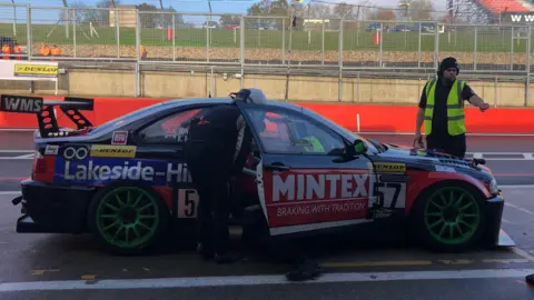 Jon Watt The back view of Jon Watt wearing overalls and a white helmet, as he bends over the open door of a BMW car which has been covered in advertisements. He is on the side of a track at Brands Hatch. Beyond him a man in a hi-vis yellow vest is pointing downwards. 
