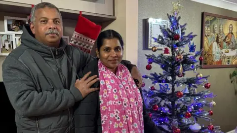 Jisha, a woman, wearing a black jacket and pink floral scarf around her neck, standing with her husband next to a purple Christmas tree.