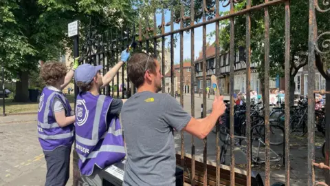 Three men working on railings outside York Minster. Two of them are wearing purple high vis jackets and one is wearing a grey t-shirt with a yellow logo on.