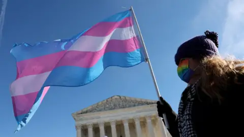  A transgender rights supporter takes part in a rally outside of the U.S. Supreme Court as the high court hears arguments in a case on transgender health rights on December 04, 2024 in Washington, DC. She is wearing a face-mask with the pride flag on it, and is waving a giant transgender flag. 