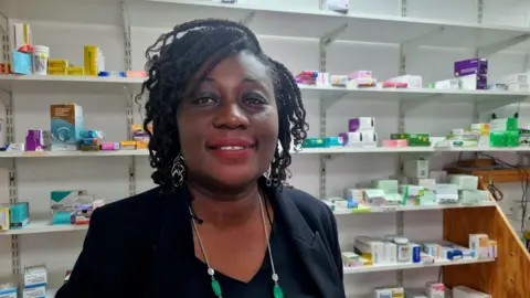 Pharmacist Irene Boateng, who has black hair and wears a smart black jacket green and silver necklace, stands in front of shelves full of medication