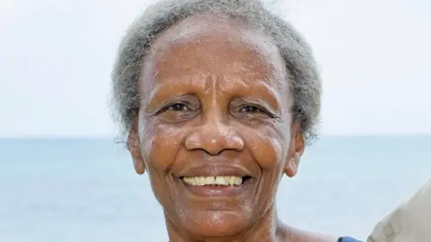 A woman with grey hair looking at the camera smiling. The sky and sea are in the background.