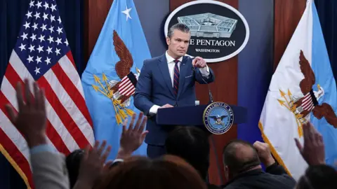 Hegseth in navy suit with red and blue tie at a podium with Pentagon seal behind him alon with flag and people raising hands in front of him