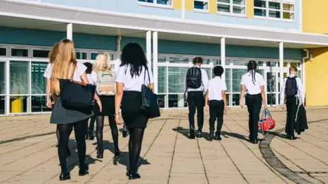An image of eight teenagers wearing school uniforms of dark skirts and tights, or trousers and white shirts walking towards a school building.