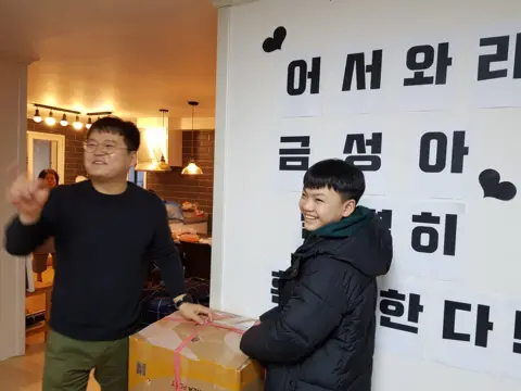 Handout A picture of a smiling teenager with boxes and his foster father, in a warm home with text behind him reading "Welcome, home Geumseong!" in Korean