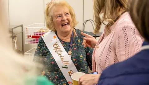 NHS Humber Health Partnership Janet Harley laughing with another woman. She has short, blonde hair and is wearing a green, floral dress and a pink and gold sash. The woman she is laughing with has long, blonde hair and is wearing a pink cardigan. She is holding a paper coffee cup and is pointing at Janet Harley.