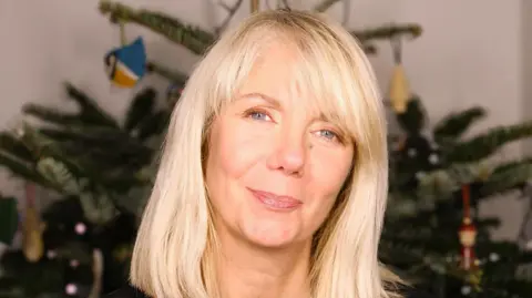 Helen Yeadon A blonde woman smiles at the camera. She is standing in front of a Christmas tree with ornaments on it.