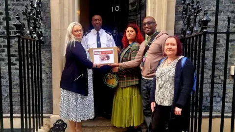 Charlotte with three members of the organisation Autistic Inclusive Meets. They are on the steps of Number 10 Downing Street. A gleaming black door is behind them and they are posing with a police officer. They are holding a cardboard box and looking at the camera.  