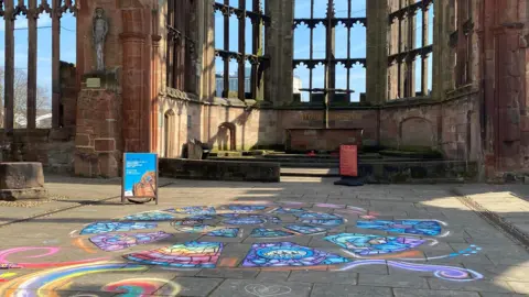 A colourful, spiralling artwork resembling a stained glass window has been created on paving slabs on the ground. The window-less frames of Coventry Cathedral's stone structure are visible behind, with blue skies.