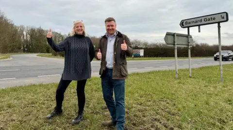 Lysette and Liam have their thumbs up and are smiling by the side of the road. The Barnard Gate sign is behind them, pointing to the left. The road to Barnard Gate comes off the A40 behind them.