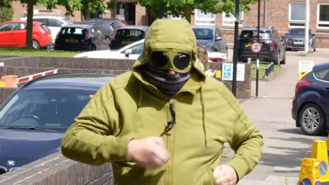 John Fairhall/BBC Man in green hoodie with attached goggles covering his eyes and a scarf over his face