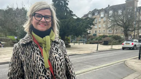 Julia Bryson/BBC A woman with blonde bobbed hair and glasses wearing a leopard print coat, green scarf and orange bag, standing on a street in Ilkley. 