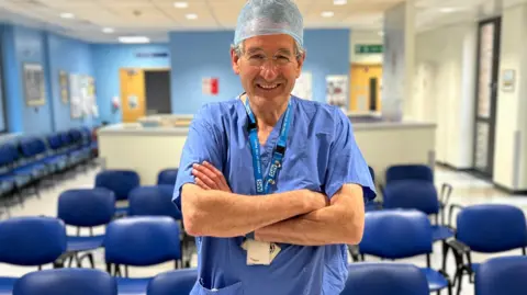 Contributed Peter Prinsley MP is smiling at the camera in a hospital waiting room. He is wearing a surgeon's blue scrubs and hat
