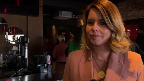 Kim McGuinness looking to the left of the camera with a slight smile. She has blonde hair which falls just above her shoulders and is wearing a large gold circular necklace and a pink blazer. She is standing in front of a bar with people lining up behind her.