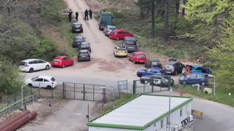 Cars parked outside the entrance to Charlton Wood. Police officers and a police vehicle are in the car park.