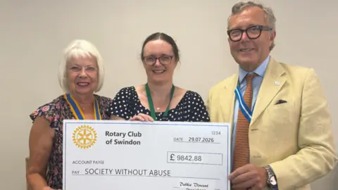 Society Without Abuse Two women and a man standing, holding a cheque for 'Society Without Abuse' for £9,842.88. They all have glasses on and are smiling.